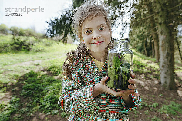 Smiling girl holding glass bottle with plants in forest