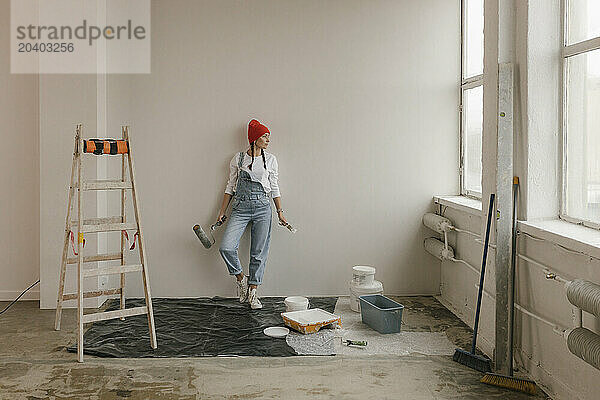 Woman leaning on white wall near ladder and paint equipment in new apartment