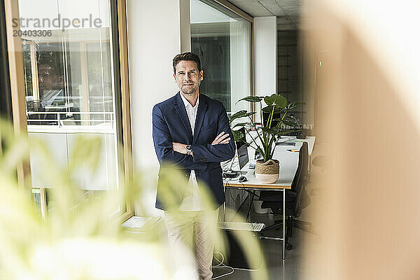 Confident mature businessman with arms crossed standing by window of office