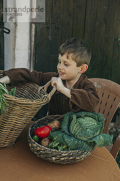 Smiling boy with vegetables in vintage wicker basket at table in backyard