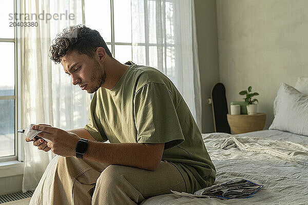 Young man calculating bills and using smart phone on bed at home