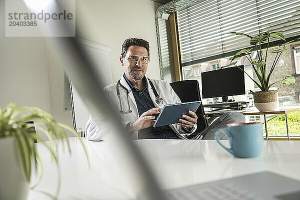 Confident doctor wearing smart glasses using tablet PC sitting at office desk in hospital