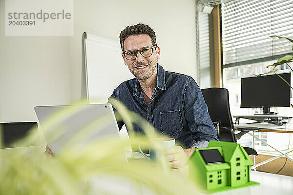 Smiling architect sitting with coffee cup and laptop by house model at desk in office