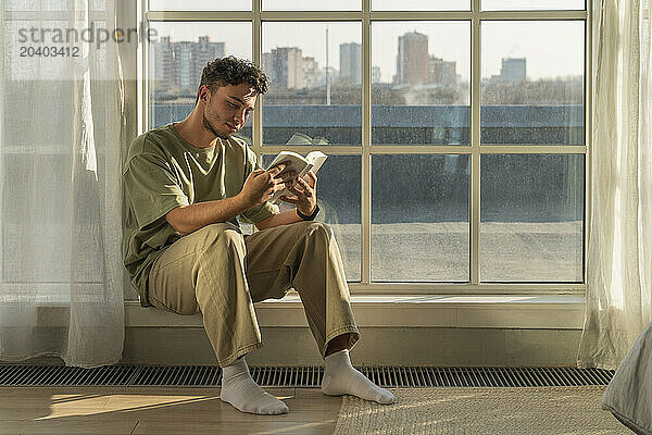 Young man sitting near window and reading book at home