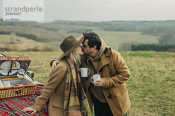 Romantic couple kissing each other and holding tea cups on picnic