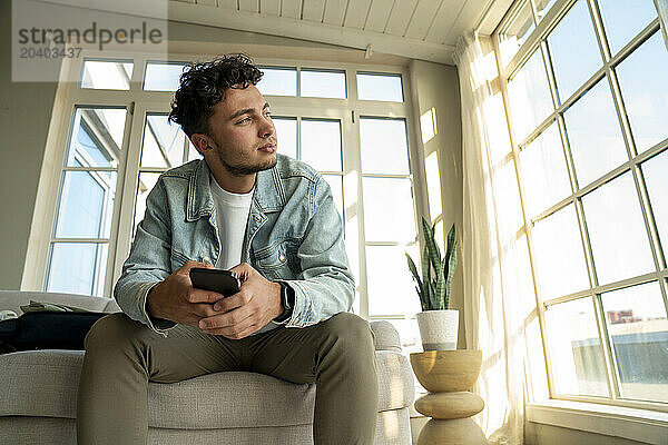 Young man sitting with smart phone on sofa at home