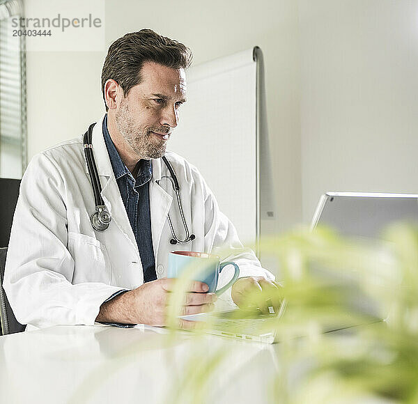 Confident doctor sitting with coffee cup using laptop at desk in office