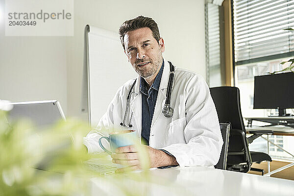 Confident doctor sitting with coffee cup and laptop at desk in office