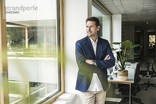 Mature businessman standing with arms crossed looking out through window of office