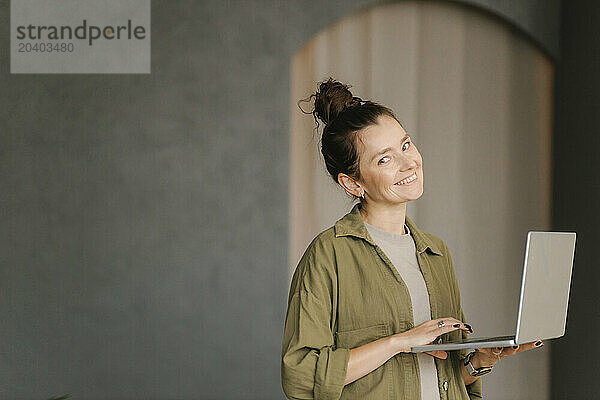 Smiling woman standing with laptop in new home