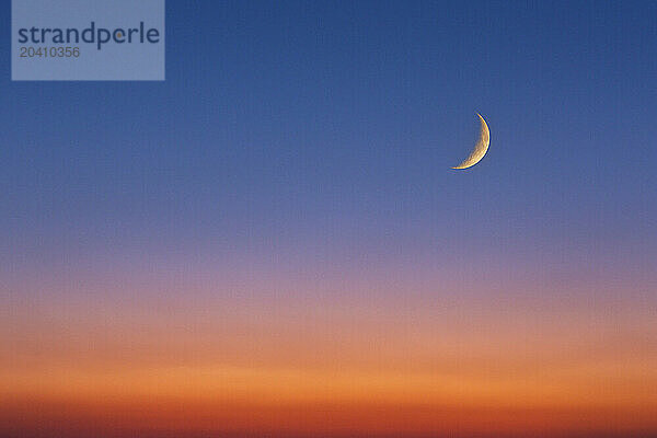 A crescent moon glowing with the warm light of sun set with a colourful glowing orange sky gradating to blue