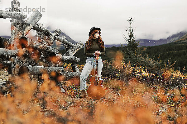 Teenage girl standing with her guitar next to a wooden fence in the fall at Glen Alps in Anchorage  Alaska