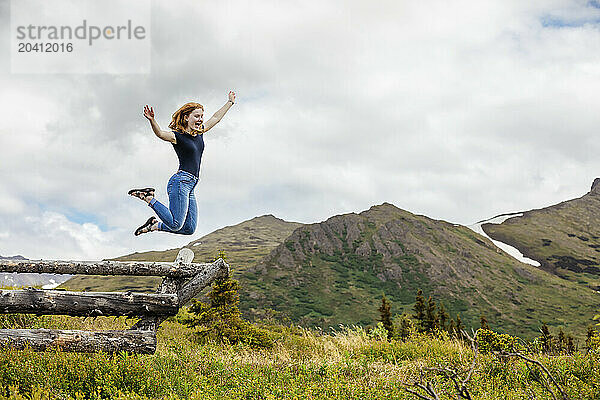 Teenage girl wearing jeans and jumping from a wooden fence  with view of the mountains.