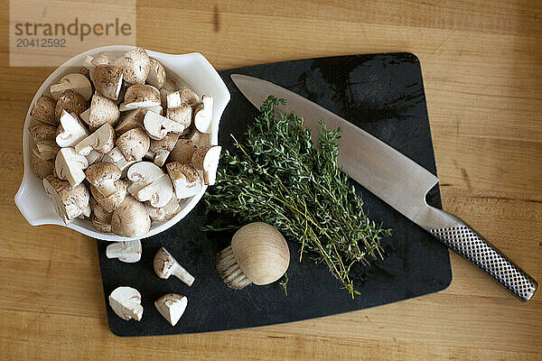 Top down view of a cutting board with cut mushrooms  a knife and some Thyme.