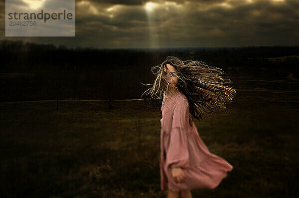 Beautiful young girl pink dress spinning in field cloudy skies