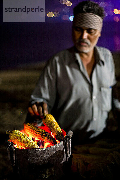 A middle aged man prepares roast-corn to sell at night on a beach.