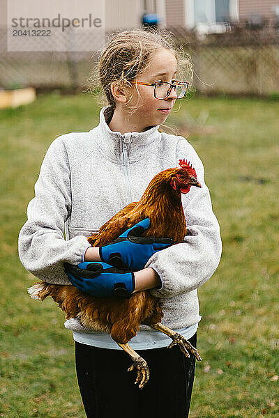 Girl with glasses holds chicken outside in yard as pet