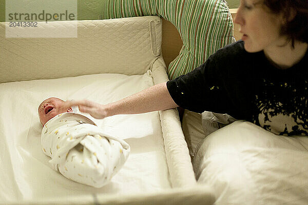 A woman soothes a swaddled crying baby in a co-sleeper crib next to her bed.