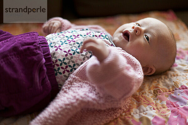 A newborn baby sits on her back on a couch smiling and looking up in a home.