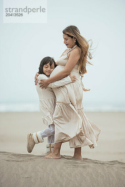 Side view of young girl hugging her pregnant mother on a cloudy beach