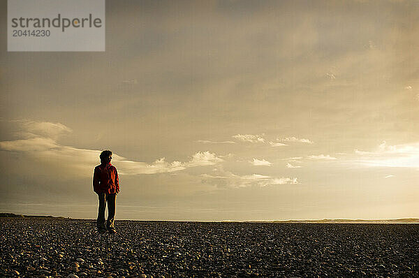 A young woman in an red jacket stands on a rocky beach at sunset in New Zealand.