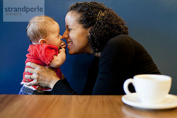 A smiling woman plays with a newborn baby in a coffee shop.