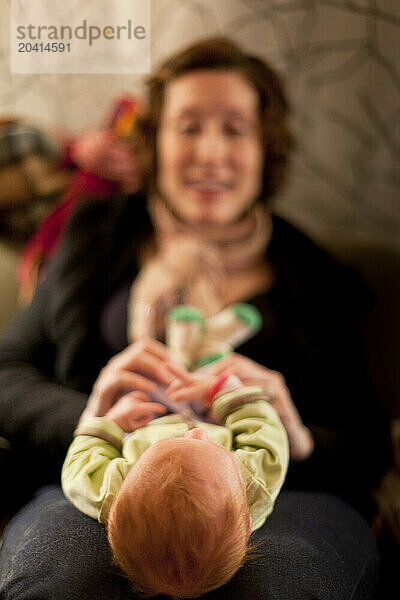 A smiling mother with red hair plays with her baby on a couch amid piles of messy laundry at night in a home.