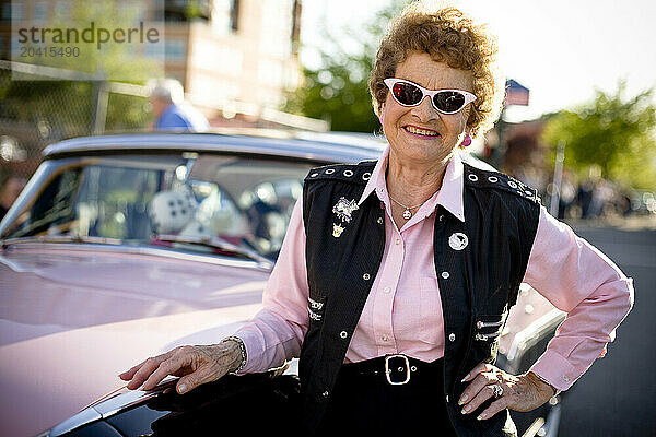 A senior aged woman in pink  poses with her classic car.