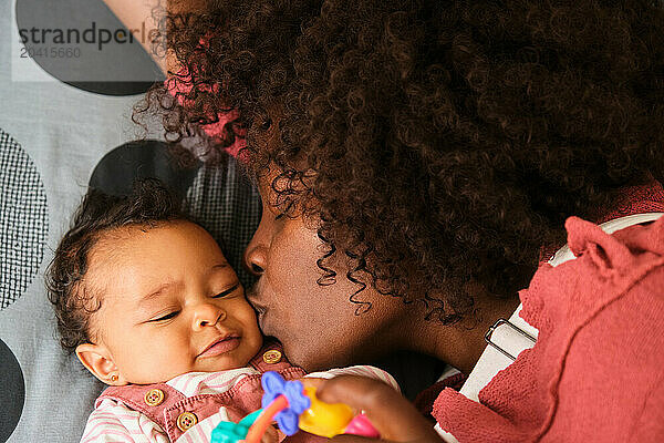 African woman is kissing a baby girl laying in bed.