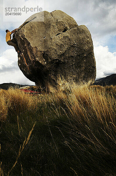 Young man climbing a boulder surrounded by tall grass and mountains in the Castle Hill Basin  New Zealand.