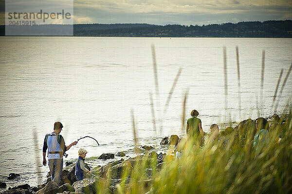 A young father carrying his baby in a sling walks with a stick over driftwood with his family on a a beach.