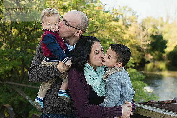 Parents kissing toddler boys on cheeks while carrying them in park