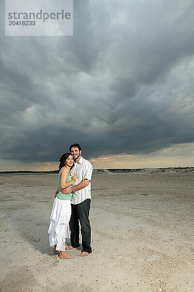 A man and a woman embrace  standing on the beach under a cloudy sky.