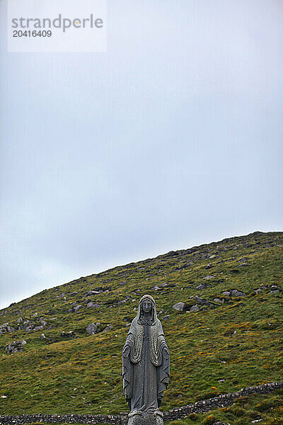 Religious statue sits in front of hill on cloudy day in Ireland.