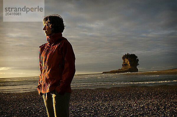 A young woman in an red jacket stands on a rocky beach at sunset in New Zealand.