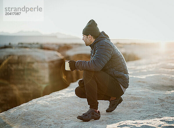 Man drinks coffee near cliff on cold morning in desert  Moab  Utah