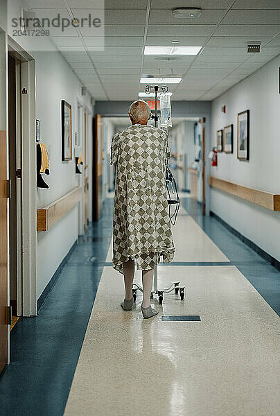 elderly male cancer patient wearing gown walks down hall in hospital