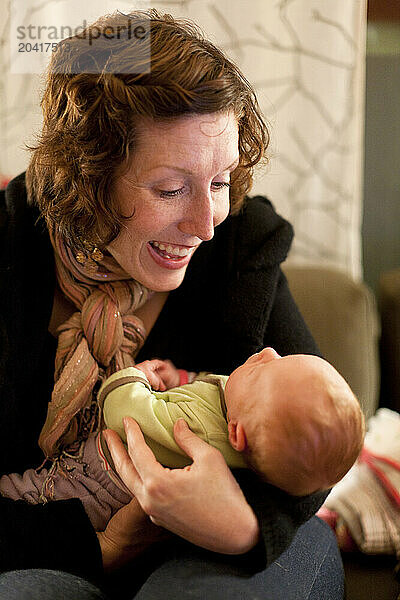 A smiling mother with red hair plays with her baby on a couch amid piles of messy laundry at night in a home.