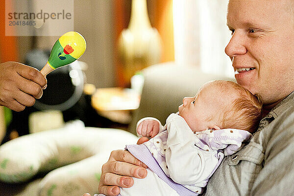 A father holds his newborn smiling baby in his arms  while a woman holds out a rattle to the baby in a living room.