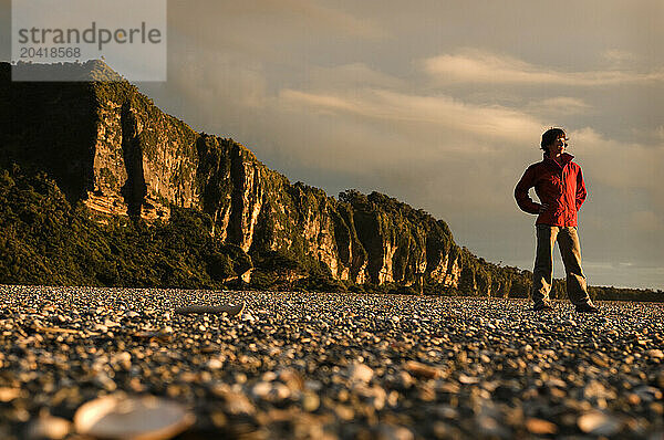 A young woman in an red jacket stands on a rocky beach at sunset in New Zealand.