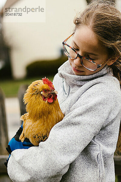 Girl in glasses holds golden chicken in yard