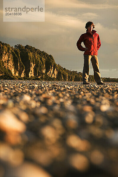 A young woman in an red jacket stands on a rocky beach at sunset in New Zealand.