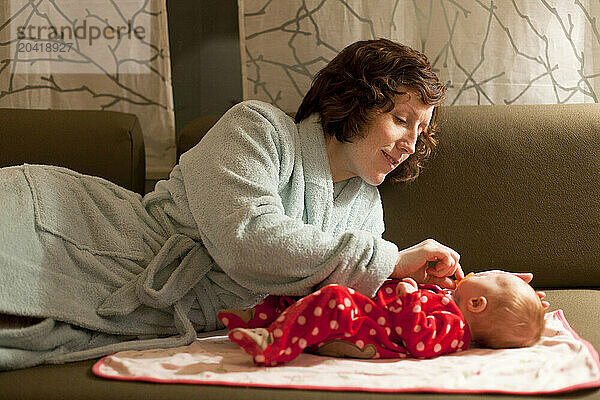 A mother in a blue bathrobe plays with her baby on a couch in her living room.