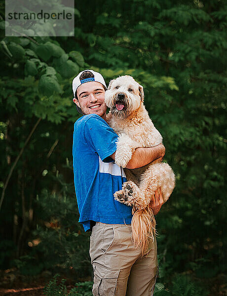 Happy older teen boy holding fluffy dog outside in park setting.