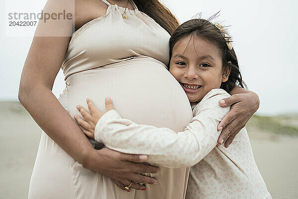 Young girl hugging her pregnant mother on a cloudy beach