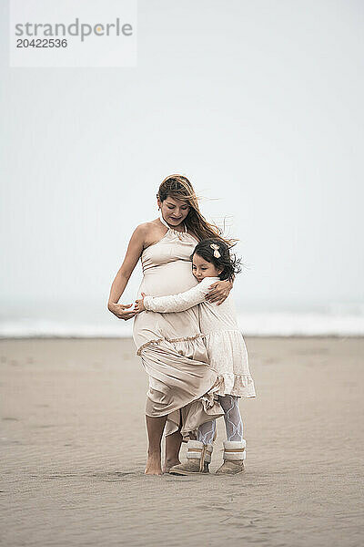 Young girl hugging her pregnant mother on a cloudy beach