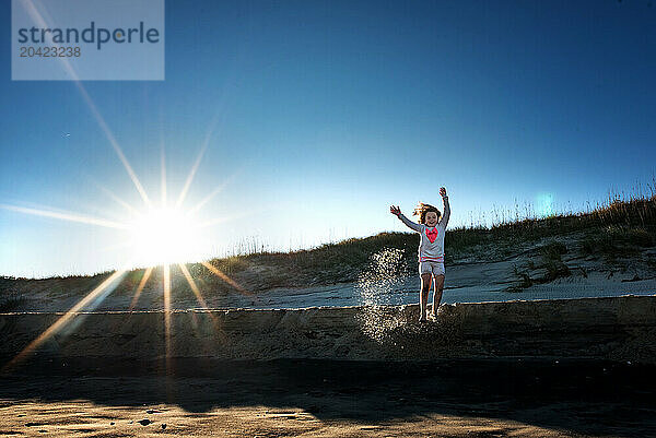 Happy tween girl jumping off sand dune with bright sunny beach