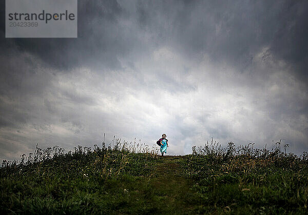 Young girl running down hill wearing purple cape under cloudy sky