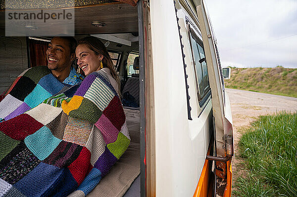 Cheerful young diverse couple in blanket sitting in travel van