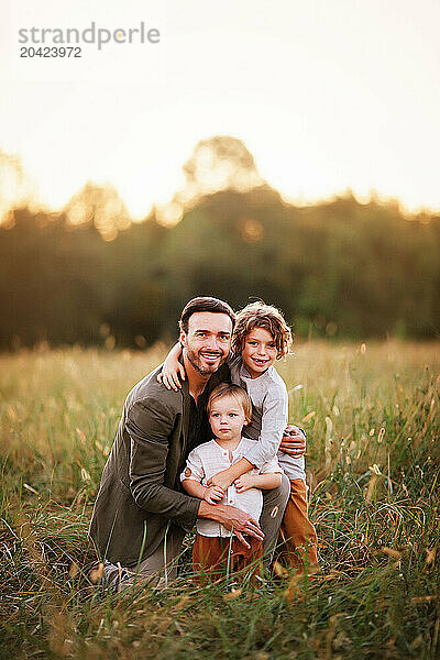 Father kneeling in a grassy field at sunset  embracing his two y